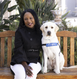 A woman sitting on a bench next to a yellow Labrador wearing a blue assistance dog vest.